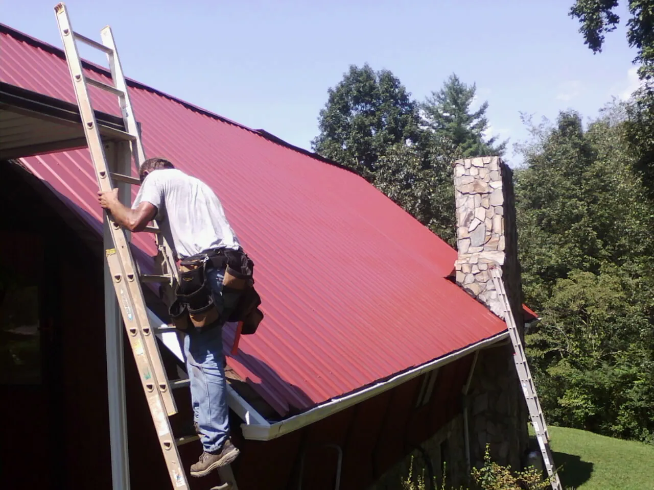 Roofer on a ladder beside a red metal roof and stone chimney — Soffit & Fascia Repair by Roofing Brothers in Crapo